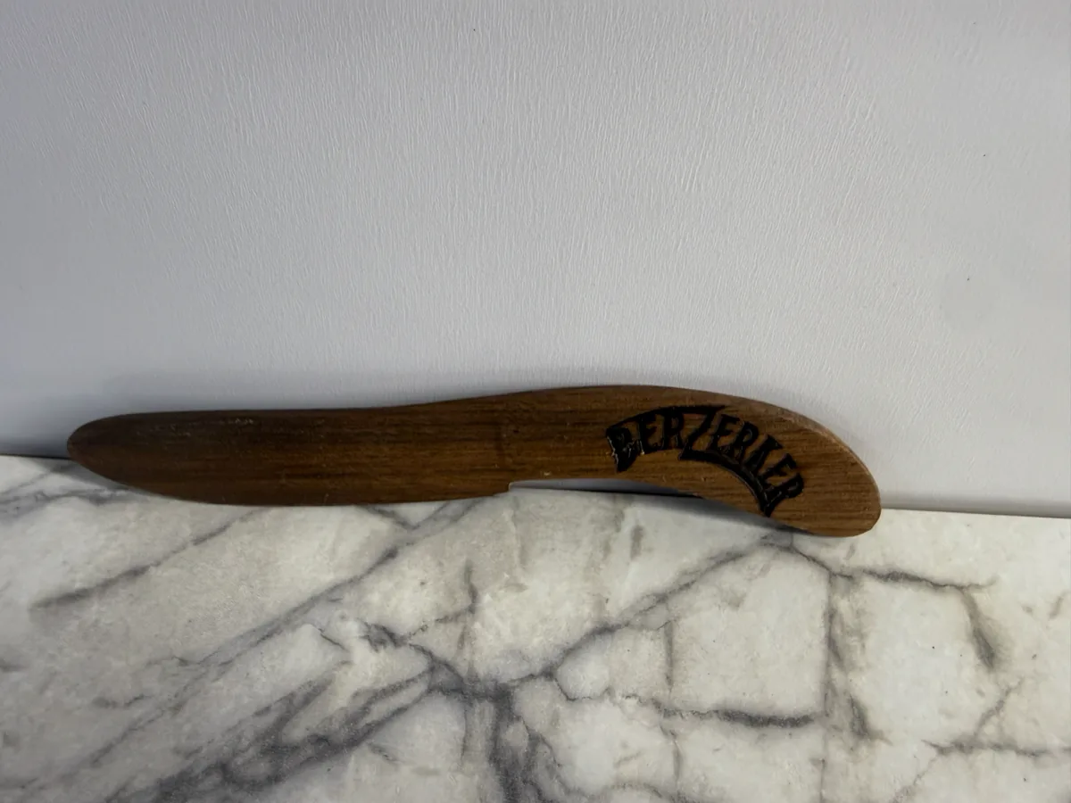 Curved wood knife sheath with a dark maker's mark on a light surface.